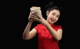 retrato asiático jovem, mulher vestido vermelho tradicional cheongsam segurando uma moeda de ouro em um saco no fundo preto foto