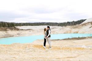 casal elegante à distância se olha e posa contra o fundo de um lago azul e areia amarela