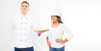 dois chefs adolescentes segurando uma placa de pizza vazia, isolada no branco. fogões americanos brancos e afro de uniforme. foto