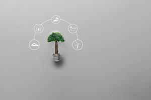 mão segurando a lâmpada com uma grande árvore em fundo cinza com fontes de energia de ícones para energias renováveis, células solares, desenvolvimento sustentável. conceito de ecologia e meio ambiente. foto