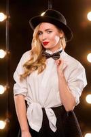 jovem loira atraente em uma camisa branca, borboleta, chapéu preto com microfone cantando no palco foto