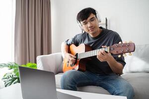 jovem bonito homem jogando acústico guitarra sentado em a sofá dentro a vivo quarto às lar. foto