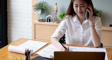 Mulher asiática sorridente atendendo uma ligação no celular durante o trabalho no laptop para o número da contabilidade financeira, relatório mensal de estatísticas de papelada