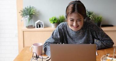 tiro recortado freelancer de mulher asiática sorridente usando fone de ouvido, comunicando-se com o cliente através de chamada de vídeo-computador. Tutora profissional agradável milenar dando aulas de idiomas online foto