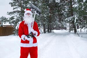 santa claus limpa neve com pá dentro inverno ao ar livre depois de uma queda de neve. limpeza a ruas dentro a Vila, compensação a passagem para carros, difícil clima condições para Natal e Novo ano foto