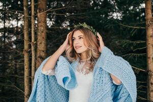 retrato do uma fofa noiva com uma guirlanda em dela cabeça, ela é embrulhado dentro uma azul cobertor contra a fundo do uma inverno floresta. inverno casamento. foto