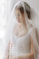 a noiva dentro uma branco vestir, coberto com uma véu, poses dentro Boa iluminação dentro dela sala. manhã do a noiva. uma lindo tiara foto