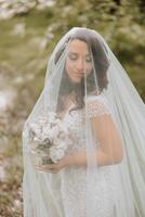 uma lindo noiva é em pé dentro uma verde Primavera floresta dentro uma glamouroso branco Casamento vestir debaixo uma véu. uma romântico lindo noiva dentro uma branco vestir é posando contra a fundo do a floresta. foto