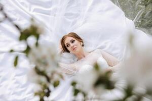 contra a fundo do uma florescendo árvore, uma ruivo noiva poses, sentado em verde grama. magnífico vestir com grandes mangas, aberto busto. Primavera Casamento foto