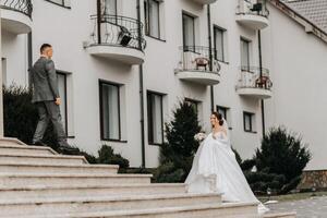 uma noiva dentro uma branco vestir com uma trem e uma noivo dentro uma terno pose em a passos do uma prédio. Casamento foto sessão dentro natureza