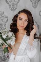 uma lindo morena noiva com uma tiara dentro dela cabelo é obtendo pronto para a Casamento dentro uma lindo roupão dentro boudoir estilo. fechar-se Casamento retrato, foto. foto