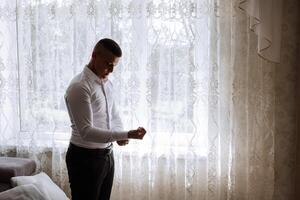 a noivo botões dele camisa de a janela. homem dentro uma camisa. masculino retrato. sessão de fotos às a janela. do noivo coleção. Casamento dia. bonito homem noivo abotoar dele camisa dentro uma hotel sala.