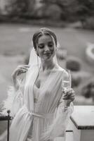 uma lindo noiva com uma grandes véu em dela cabelo, vestindo a elegante branco penhoar, em pé em uma sacada negligenciar a cidade, bebendo uma beber durante a Casamento manhã. Preto e branco foto