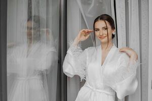 retrato do noiva menina dentro branco vestido com profissional Penteado e natural Maquiagem dentro hotel quarto com reflexão dentro janela. a melhor dia foto