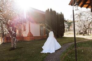 a noiva tem dela ombros virou para a Câmera, caminhando ao longo uma pedra caminho. uma grandes trem. magnífico vestir com grandes mangas, aberto busto. Primavera Casamento foto