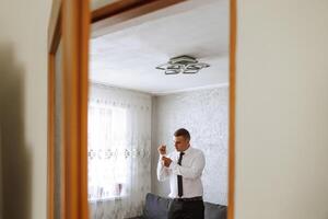 a noivo botões dele camisa de a janela. homem dentro uma camisa. masculino retrato. sessão de fotos às a janela. do noivo coleção. Casamento dia. bonito homem noivo abotoar dele camisa dentro uma hotel sala.