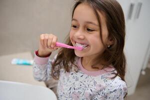 adorável pequeno criança menina escovar dentes dentro frente do a espelho dentro a casa banheiro foto