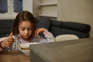 adorável pequeno criança menina levando dela saudável café da manhã às lar, comendo saudável flocos com vegetariano leite foto