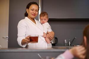 feliz sorridente mulher, jovem mãe carregando dela bebê garoto, segurando uma copo do quente chá enquanto em pé às cozinha contador foto