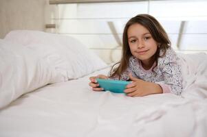 pequeno criança menina dentro pijamas, deitado em a cama, segurando Móvel telefone e jogando conectados vídeo jogos, sorridente às Câmera foto