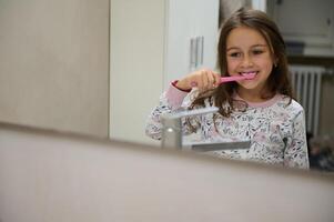 adorável pequeno criança menina olhando às dela espelho reflexão enquanto escovar dentes dentro a a banheiro. dental Cuidado higiene foto