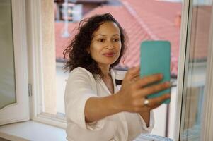 bonita mulher em pé às janela negligenciar paisagem urbana com vermelho cobertura azulejos em casas, sorrisos levando selfie em Smartphone foto