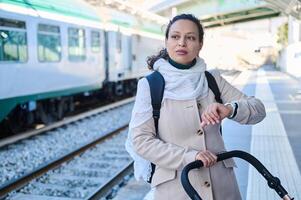 jovem mãe empurrando uma bebê carrinho de bebê, verificação Tempo em relógio de pulso enquanto esperando a trem em a estrada de ferro estação plataforma foto