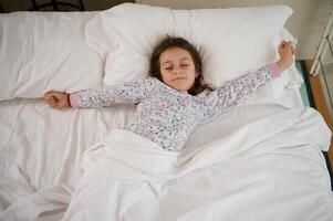 Visão a partir de acima do uma pequeno menina deitado em a cama e alongamento dela braços para cima enquanto acordando acima dentro a manhã foto