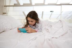 Internet vício dentro infância. pequeno menina tendo Diversão em Smartphone enquanto deitado em cama. a conceito do digital aparelhos foto