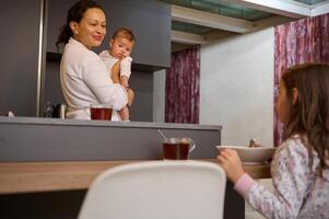 alegre mãe segurando dela bebê e desfrutando levando chá com dela filha dentro a casa cozinha dentro a manhã foto