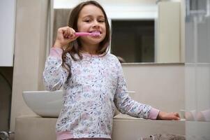 pequeno criança menina escovar dela dentes, em pé dentro a cinzento minimalista casa banheiro, sorridente olhando às Câmera. oral Cuidado foto