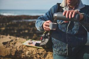 detalhes em garrafa térmica frasco dentro a mãos do sorridente turista caminhante derramando alguns chá para dentro aço caneca, sentado em uma Rocha de mar foto