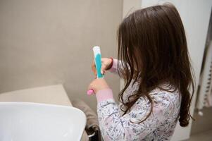 caucasiano pequeno criança menina apertando pasta de dentes em dela Rosa escova de dente, em pé às branco lavatório dentro a casa banheiro foto