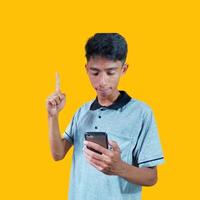 jovem exterior olhando ásia homem vestindo cinzento camiseta, segurando índice dedo inteligente telefone com Novo ise, isolado em amarelo fundo foto