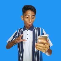 jovem ásia homem surpreso olhando às inteligente telefone, vestindo colorida camiseta, isolado azul fundo. foto