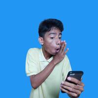 jovem ásia homem surpreso olhando às inteligente telefone, vestindo amarelo camiseta, isolado azul fundo. foto