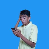 jovem ásia homem surpreso olhando às inteligente telefone, vestindo amarelo camiseta, isolado azul fundo. foto