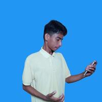 jovem ásia homem surpreso olhando às inteligente telefone isolado azul fundo. vestindo uma amarelo camiseta foto