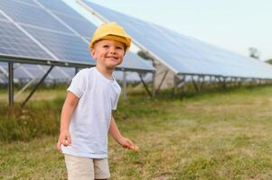 uma feliz pequeno Garoto dentro uma amarelo capacete é em pé em uma solar painel Fazenda. foto