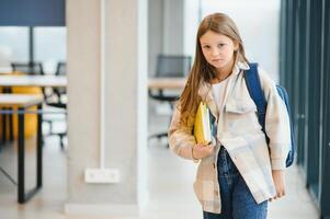 pequeno lindo escola menina em pé entre corredor às escola, segurando notas às mãos. engraçado e feliz menina sorridente às Câmera, em repouso depois de lições em primário escola foto