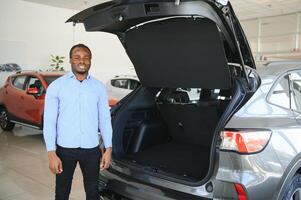 feliz carro comprador, Novo carro proprietário conceito. retrato do animado jovem africano americano cara dentro concessionária sala de exposições foto