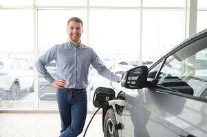 barba homem tentando uma Novo cobrando cabo com uma carro cobrando estação às a motor concessionária. conceito do comprando elétrico veículo. inteligente ecológico vivo foto