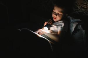 Garoto com lanterna lendo livro debaixo cobertor às lar. foto