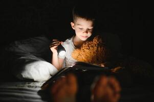 criança lendo livro dentro cama. crianças ler às noite. pequeno Garoto com fada conto livros dentro quarto . Educação para jovem crianças. hora de dormir história dentro a tarde. fofa criança debaixo cobertor dentro Sombrio quarto com luminária foto