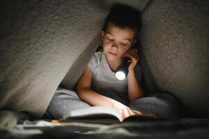 criança lendo livro dentro cama. crianças ler às noite. pequeno Garoto com fada conto livros dentro quarto . Educação para jovem crianças. hora de dormir história dentro a tarde. fofa criança debaixo cobertor dentro Sombrio quarto com luminária foto