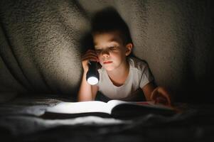 Garoto com lanterna lendo livro debaixo cobertor às lar. foto