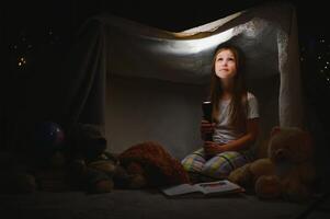 feliz criança menina rindo e lendo livro dentro Sombrio dentro uma barraca às lar. foto