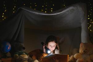 pequeno menina estão lendo uma livro com lanternas dentro barraca. feliz menina jogando às lar. engraçado adorável criança tendo Diversão dentro quarto foto