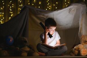 Garoto lendo livro com lanterna dentro barraca às noite. foto