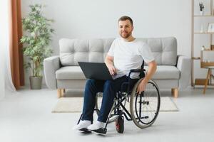 retrato do sorridente pessoa com uma incapacidade sentado dentro cadeira de rodas e trabalhando em computador portátil a partir de lar. foto